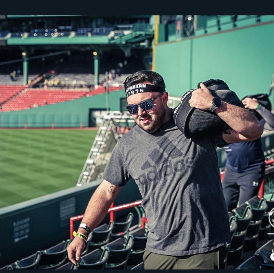 Tyler Moore — Day 12 of the 100 in 100 Member Stories series at Prototype Training Systems. Tyler is photographed at Fenway Park during a Spartan race, carrying a sandbag on his shoulders. Member since 2023, 500 sign-ins, 415 lb deadlift.